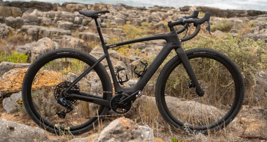 A matte-black bicycle, Specialized Turbo Creo 2 e-bike, touted as the lightest electric bike, stands proudly on a rocky terrain with knobby tires gripping the uneven ground. In the background, shrubs and a serene water body complete this adventurous scene.