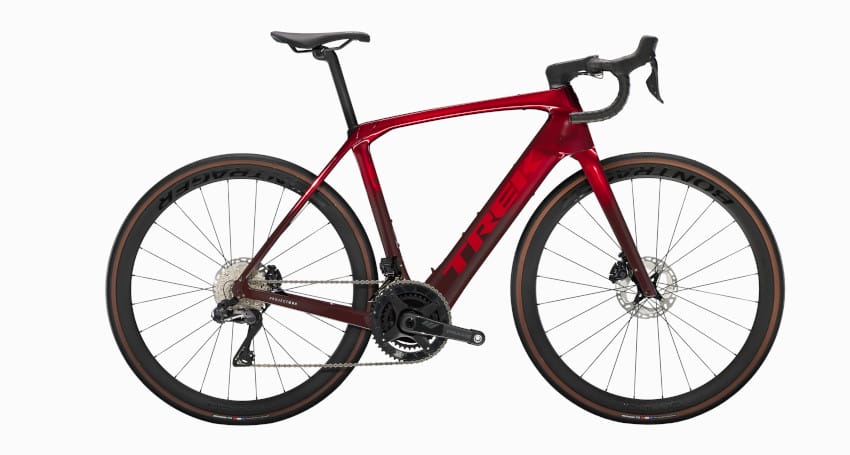 A red Trek Domane road bicycle with drop handlebars, thin tires, and a carbon fiber frame. The bike, considered one of the lightest electric bikes on the market, features a disc brake system and is equipped with a Shimano gearset.