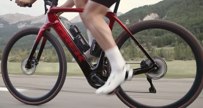 Close-up of a cyclist pedaling the lightest electric bike, a Trek Domane e-bike, painted a sleek red, with a mountainous landscape in the background.