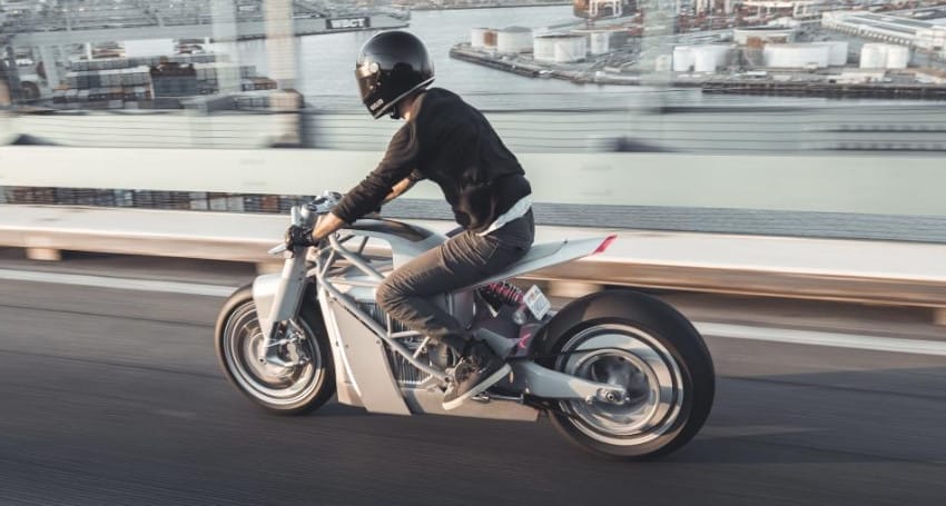 A person wearing a black helmet rides a sleek, modern electric motorbike on an elevated roadway with an industrial waterfront in the background, capturing the essence of Europe's evolving urban landscape.