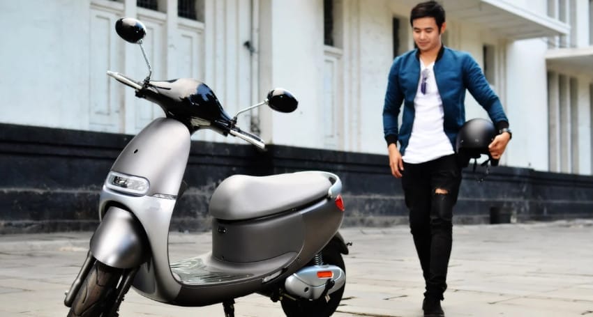 A person in a blue jacket and ripped black jeans is holding a helmet, standing near a parked grey scooter—typical of the electric motorbikes popular in Indonesia's urban settings.