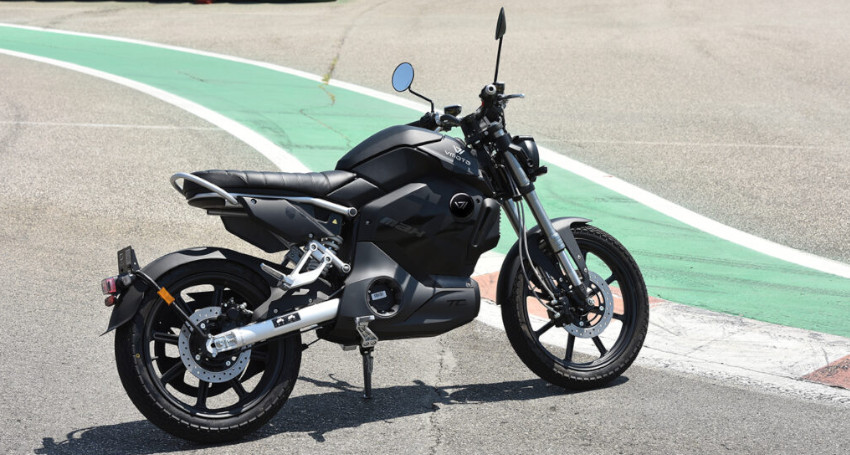 A modern black motorcycle with a superfast charging battery is parked on an asphalt road next to a green and white painted area. The bike has a sleek design with one mirror and an angled stand.
