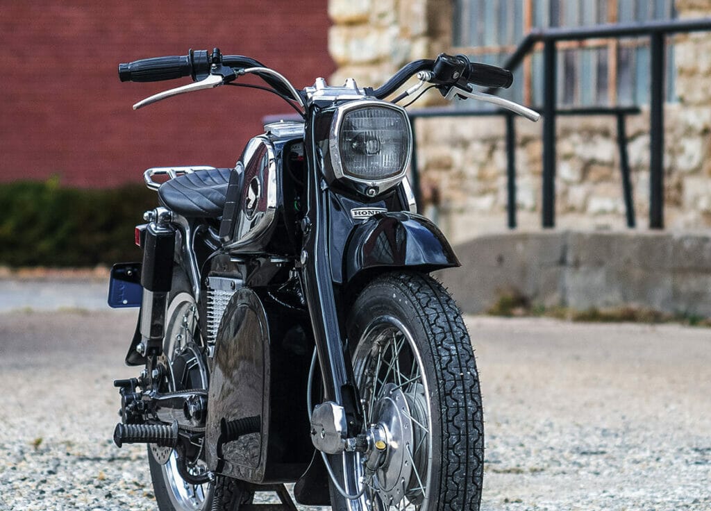 1967 Honda CA160 Dream Motorcycle transformed into a seamless and silent beauty, parked in front of a brick building.