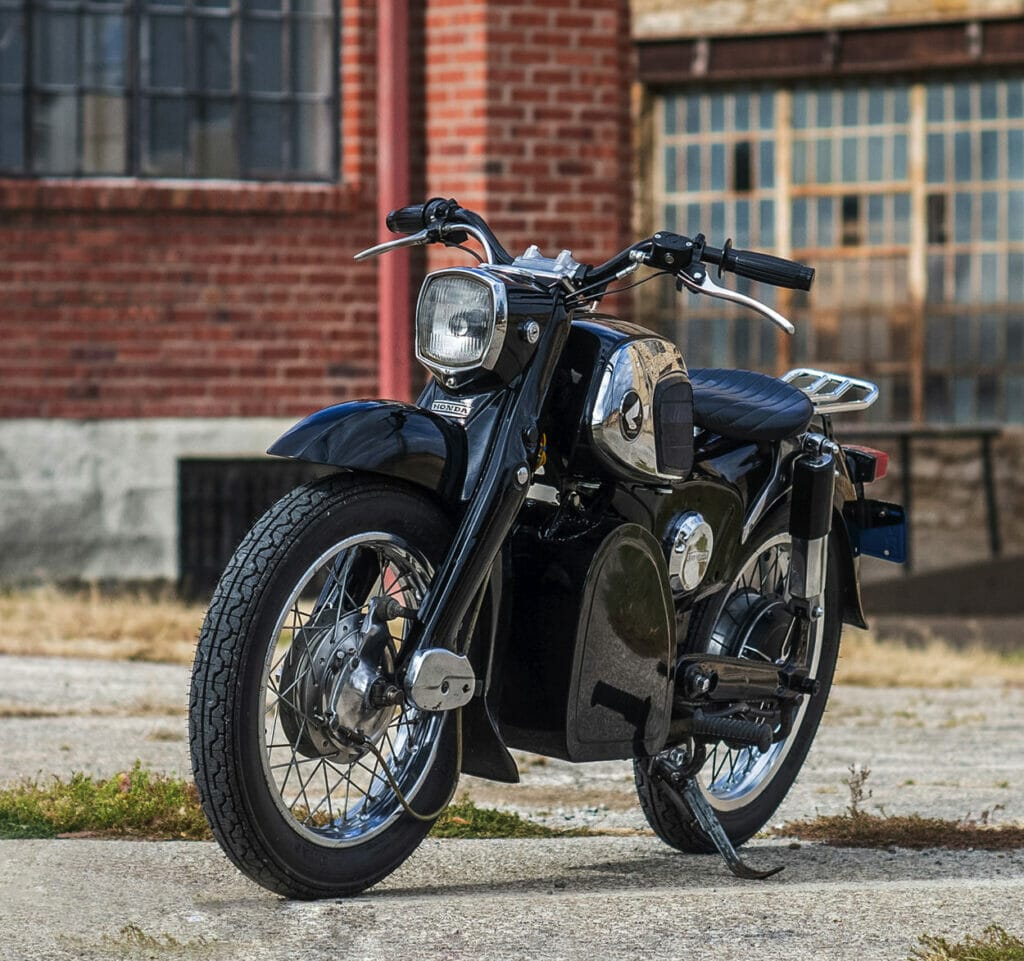 A silent black motorcycle, the 1967 Honda CA160 Dream, seamlessly converted into an electric beauty, parked in front of a brick building.