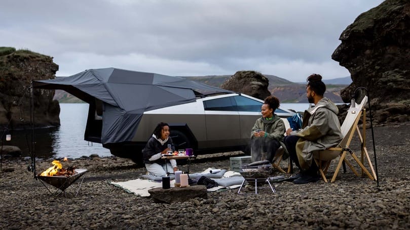 A group of people sitting around a campfire next to a (Tesla) car.