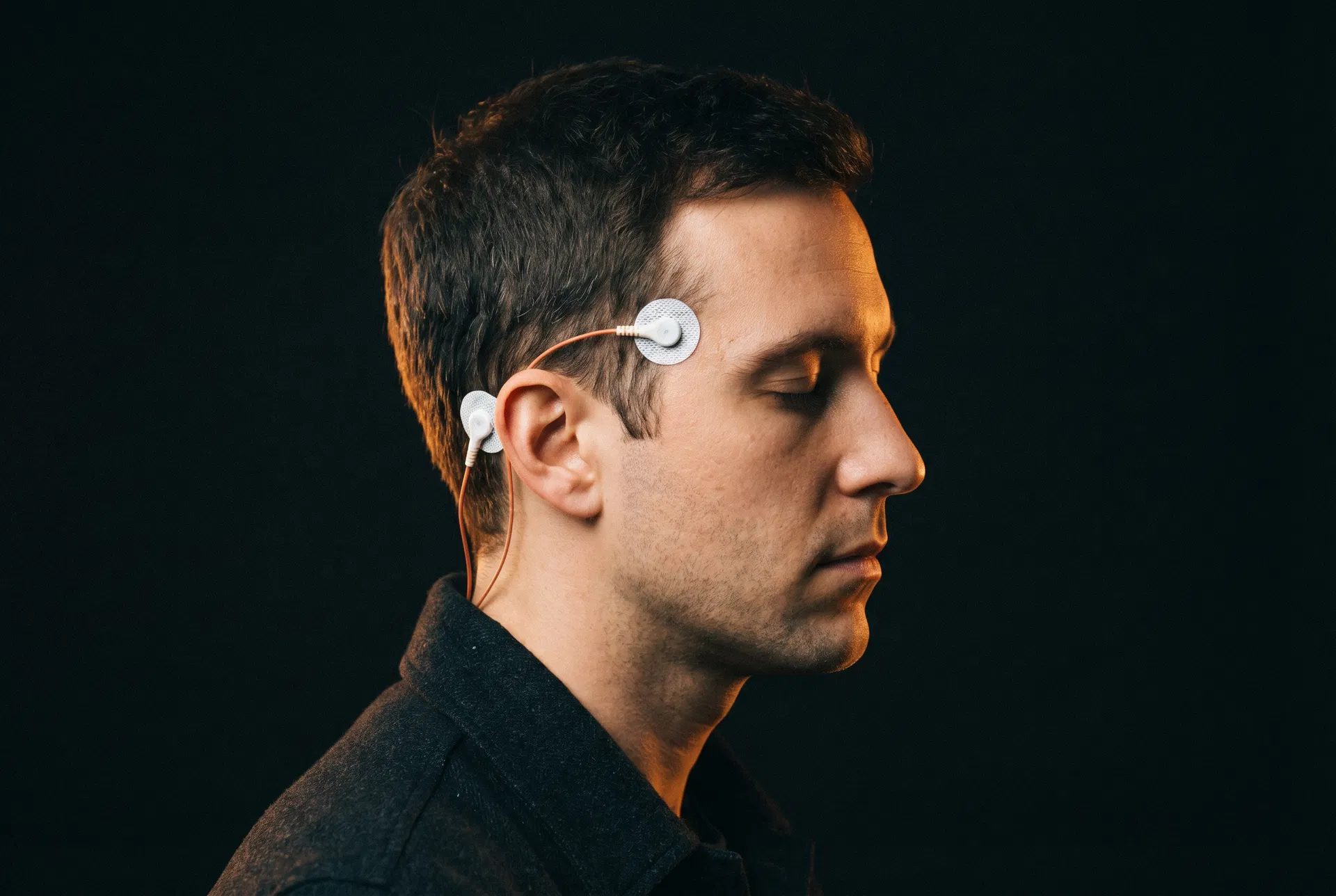 Editorial close-up of a young man with two clinical electrodes on his temple, eyes closed, against a deep ink background.