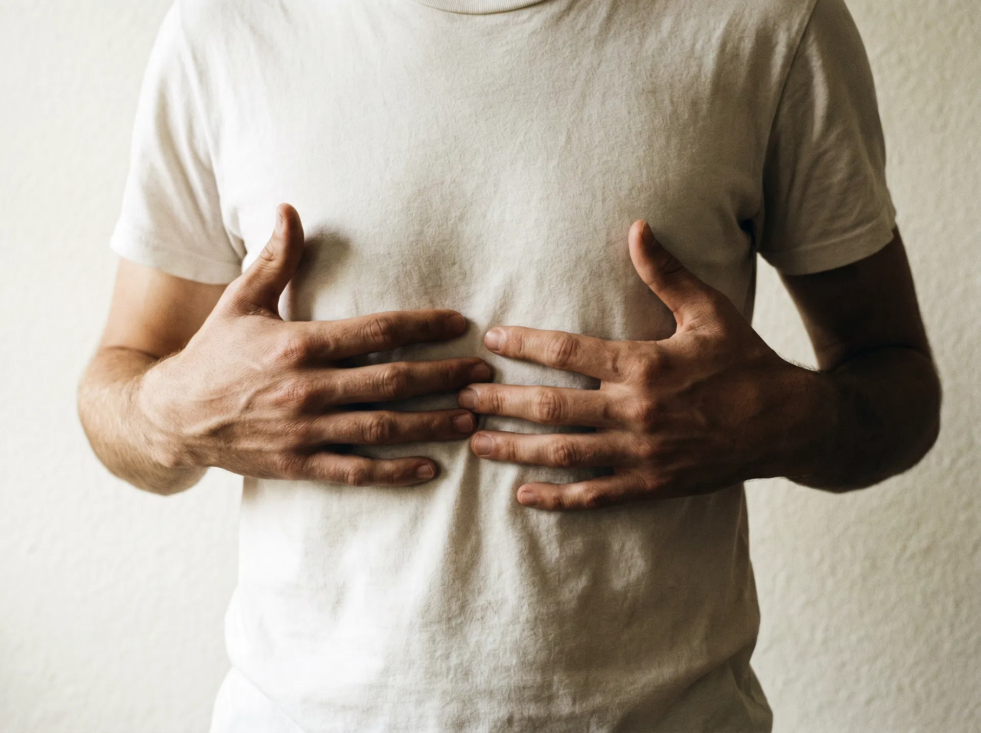 A young man's hands rest open on his diaphragm in a plain white t-shirt, ribcage softly visible in window light.
