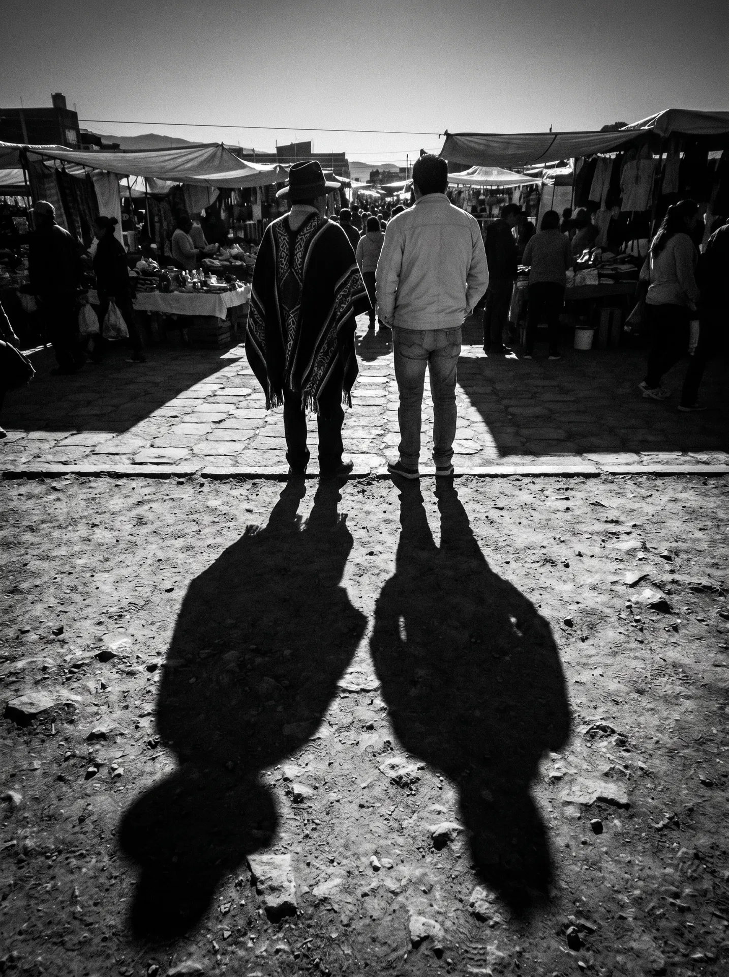 Two figures at the edge of a market — one in traditional woven garment, one in modern dress. They face the same direction, standing close enough that their shadows merge on the stone ground. The shadow merger is the subject. Fuji X-Pro1 aesthetic, Graciela Iturbide observational lineage.