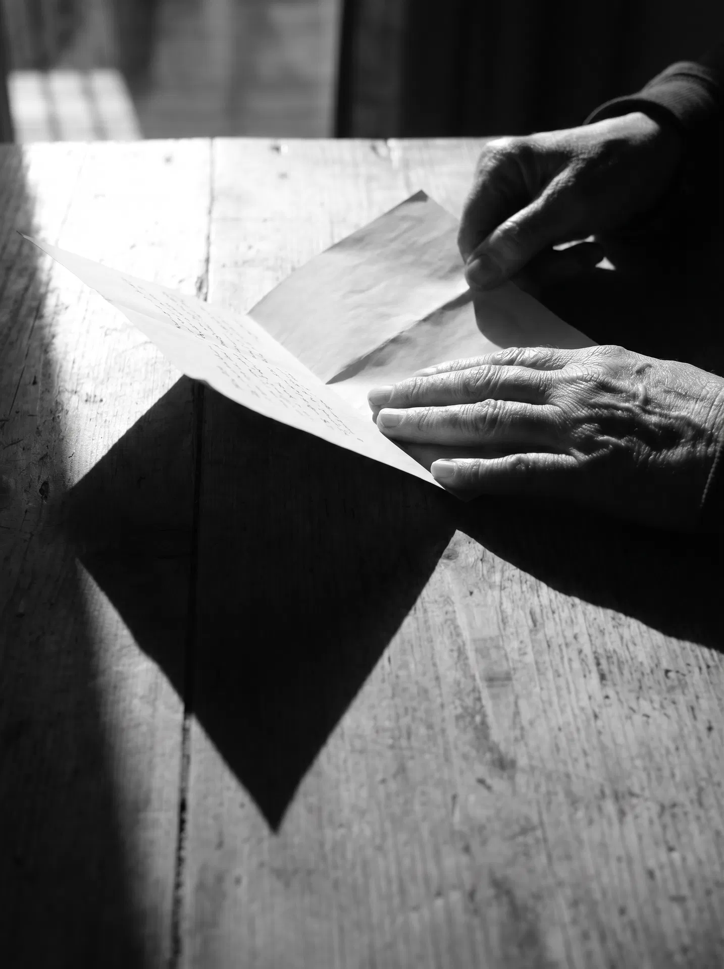 A woman's hands place a single letter on a bare table. The hands are the entire subject. The table is empty except for this one placed thing. The gesture is deliberate, unhurried, complete. Fuji X-Pro1 aesthetic, Carrie Mae Weems observational lineage.