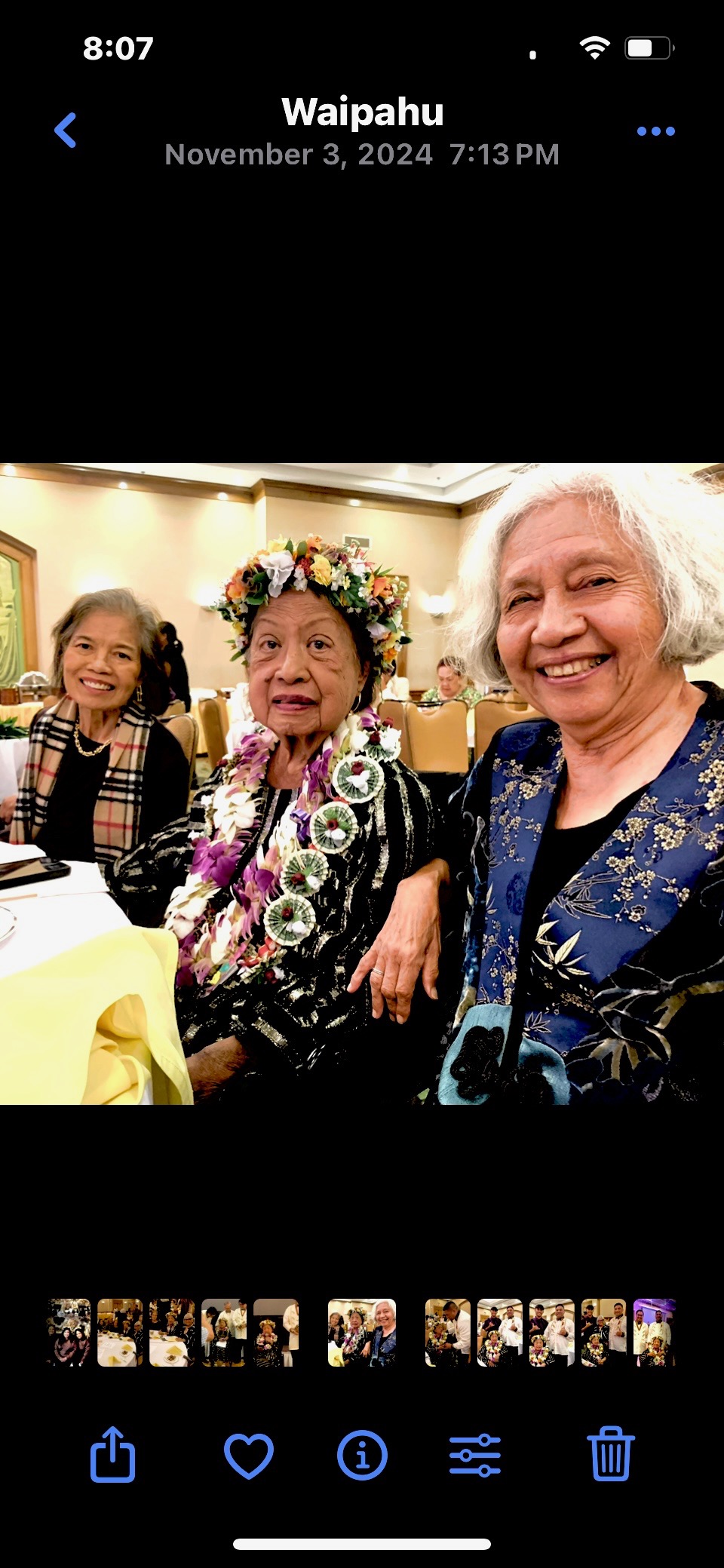 Three women seated at a banquet table during a celebration in Waipahu, Hawaii, November 2024. The woman in the center wears a colorful flower haku lei crown and a money lei, suggesting she is the guest of honor. She appears to be an elderly Filipino woman. The women beside her are smiling warmly.
