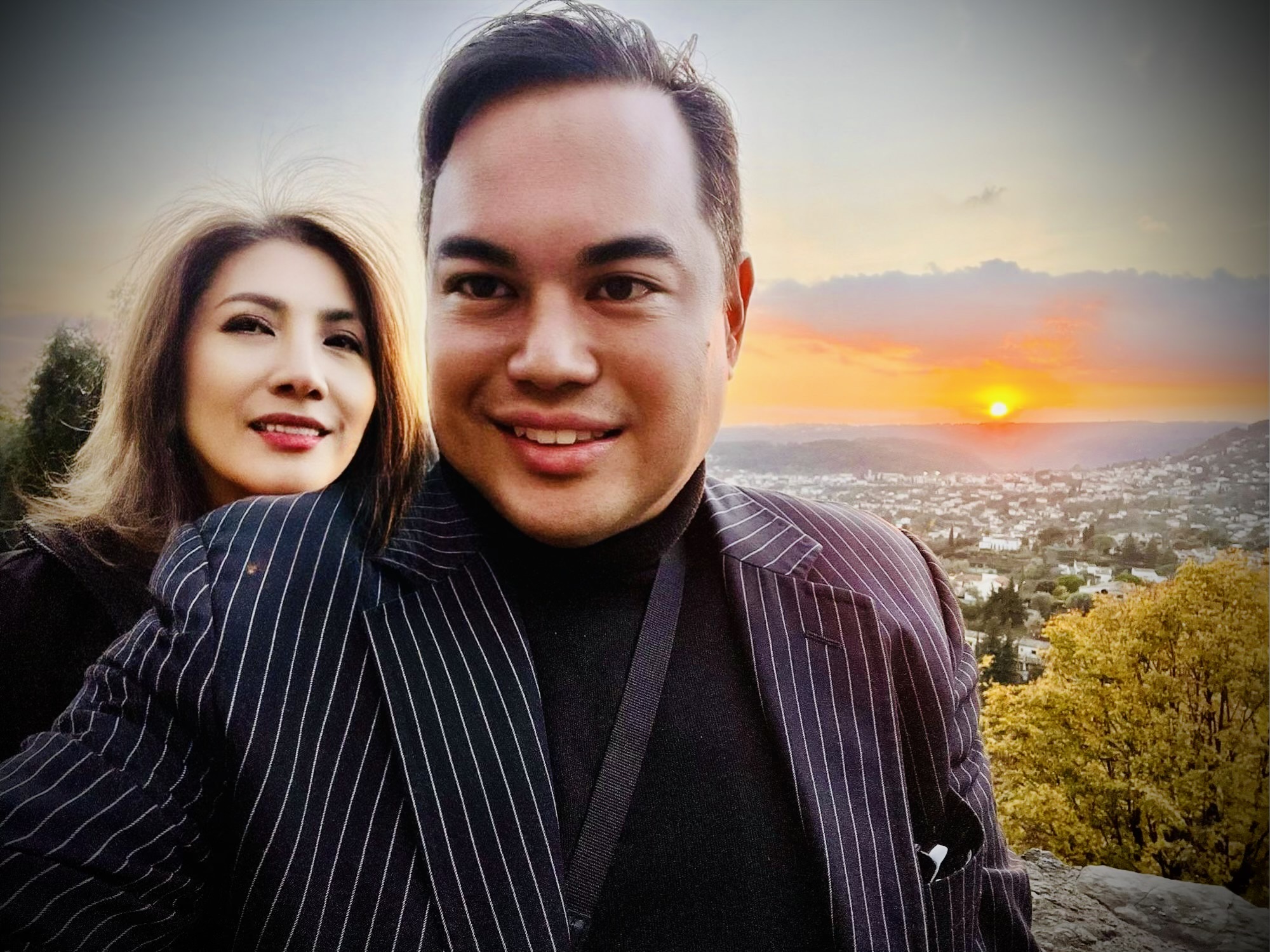 A couple posing for a selfie at sunset with a panoramic view of a city below. The woman has highlighted hair and is wearing a dark pinstripe blazer. The man is wearing a matching pinstripe suit with a black turtleneck. The golden sunset and cityscape create a warm, romantic backdrop.