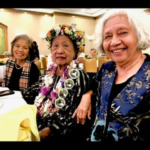 Three older women are posing for a photo at an indoor event. The woman in the center is wearing a flower crown and several leis, suggesting she is being celebrated. The women on either side of her are smiling warmly at the camera.