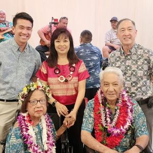 A multi-generational group of people are posing for a photo at an indoor event. An elderly couple is seated in the front, both wearing leis, with the woman also wearing a floral head wreath. Three younger adults stand behind them, and a band with several musicians is visible in the background, creating a warm and celebratory atmosphere.