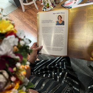 An older person is seen holding a book or publication open to a page titled 'Milagros Medallon Kaahanui'. The page contains a photograph of Ms. Kaahanui and a detailed biography of her life and work. The scene is intimate and appears to be indoors, with a bouquet of flowers in the foreground, suggesting a moment of quiet remembrance or study of her legacy.