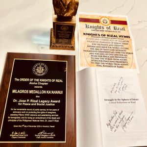 The image displays a collection of items related to an award. There is a wooden plaque from The Order of the Knights of Rizal, a golden bust trophy, a paper with the Knights of Rizal Hymn, and an open book with a handwritten message. The award, the Dr. Jose P. Rizal Legacy Award for Peace and Social Justice, was presented to Milagros Medallon Ka'ahanui.
