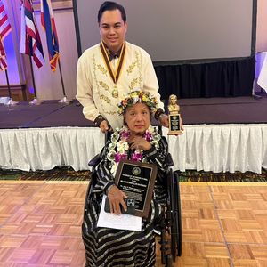 A woman in a wheelchair is receiving an award from a man standing behind her. The woman is wearing a black and gold dress, a floral headpiece, and a lei. She is holding a plaque. The man is wearing a traditional Filipino Barong Tagalog. They are on a stage in front of a screen, with flags in the background.
