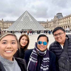 A family of four is taking a selfie in front of the Louvre Museum in Paris. The iconic glass pyramid is visible in the background. The family appears to be on vacation and are smiling for the camera.
