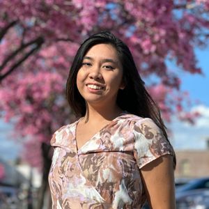 A young woman with long, dark hair is smiling warmly at the camera. She is wearing a patterned dress and is standing outside with a beautiful background of a tree with vibrant pink blossoms, which are slightly out of focus. The lighting is bright and sunny, highlighting her features.