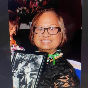 A smiling woman with glasses and a black lace top is holding a framed black and white photograph of another person. The scene appears to be an indoor event, with floral arrangements visible in the background.