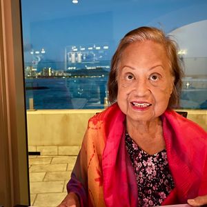 An elderly woman with a warm smile is seated at a restaurant table. She is wearing a vibrant, multi-colored scarf over a black and pink floral top. Behind her, a large window reveals a city skyline across the water at dusk. An open menu sits on the table in front of her.