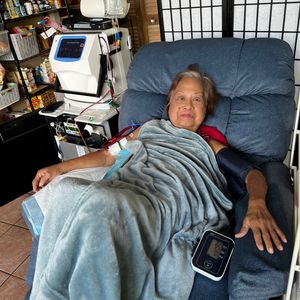 An elderly woman is seated in a large, comfortable-looking blue recliner, covered with a light blue blanket. She is undergoing a medical treatment, likely home dialysis, as indicated by the medical equipment next to her, including a dialysis machine and IV drip. A blood pressure cuff is on her arm, with the monitor resting on the chair's armrest.