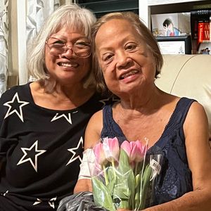 Two older women are sitting next to each other on a couch and smiling at the camera. The woman on the right is holding a bouquet of pink tulips. They appear to be in a cozy, indoor setting, possibly a living room, with a bookshelf visible in the background.