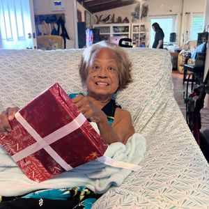 An elderly woman with white hair is sitting up in a couch or bed, smiling warmly as she holds a red and white wrapped present. She is covered by a light blue blanket, and the background appears to be a cozy, lived-in home environment. Another person can be seen in the background, out of focus.