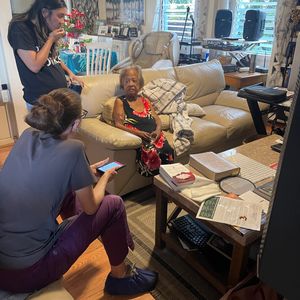 An elderly woman is sitting on a couch while two younger women are with her in a home setting. One of the younger women is wearing scrubs and looking at a phone, while the other is standing and talking to the elderly woman. The scene appears to be a home visit or a casual gathering.