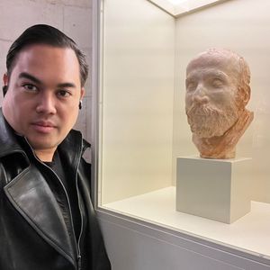 A person with dark hair, wearing a black leather jacket, takes a selfie next to a terracotta bust of a bearded man. The bust is displayed inside a glass case on a white pedestal in what appears to be a museum or gallery.