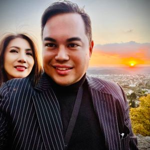 A man and a woman are taking a selfie with a scenic sunset in the background. The man is in the foreground, wearing a pinstripe jacket over a black turtleneck. The woman is behind him, smiling. The sun is setting over a town or city in the distance, casting a warm glow over the scene.