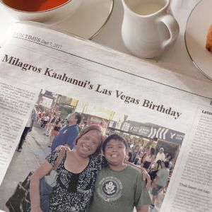 The image shows a newspaper titled 'FUN TIMES' from September 7, 2017. The headline of the newspaper is 'Milagros Kaahanui's Las Vegas Birthday' and it features a photo of a woman and a young boy smiling together. The newspaper is laid out on a table next to a cup of tea, a small pitcher of milk, and a pastry.