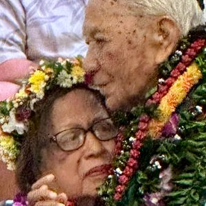 An elderly man with white hair affectionately kisses the forehead of an elderly woman with dark hair and glasses. Both are adorned with beautiful, colorful leis, suggesting a special occasion or celebration, likely in a Hawaiian context. The image captures a tender, intimate moment between the two individuals.