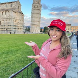 A young woman with a red 'MAKE AMERICA GREAT AGAIN' hat is posing in front of the Leaning Tower of Pisa. She is smiling and wearing a pink jacket. The image is taken outdoors in a grassy area with the tower and another building in the background.