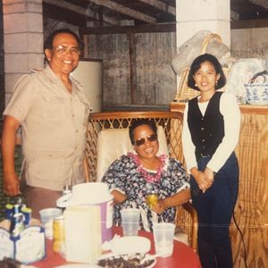 The image captures a warm, informal gathering of three people. A smiling man in a safari-style shirt stands next to a seated woman wearing sunglasses and a floral dress with a lei. Another woman in a vest and long-sleeved shirt stands beside them, creating a friendly, familial scene around a table with food and drinks.