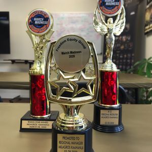 This is an image of three trophies on a wooden desk. The trophies are from an event called 'March Madness 2020' by 'Freedom Equity Group'. The awards are for 'Rising Star Award', 'Promoted to Regional Manager', and '#23 Personal Recruiter', all awarded to Milagros Kaahanui on March 28, 2020.