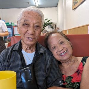 The image shows a heartwarming portrait of an elderly couple seated together in what appears to be a restaurant. The man, with white hair and a gentle smile, looks directly at the camera, while the woman rests her head on his shoulder with a joyful expression. Their closeness and relaxed posture suggest a deep, affectionate bond.