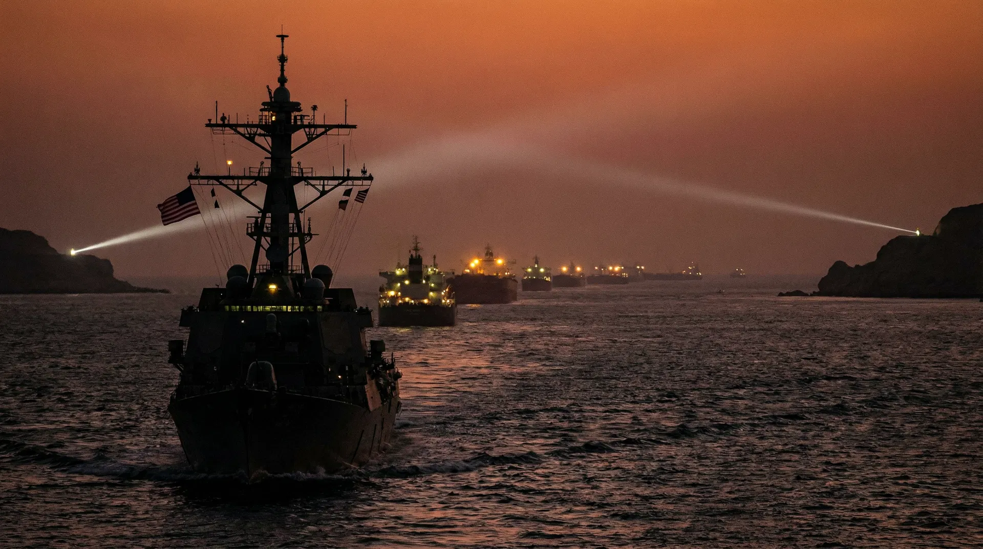 A US Navy ship patrols the Strait of Hormuz at dusk, with oil tankers in the background.