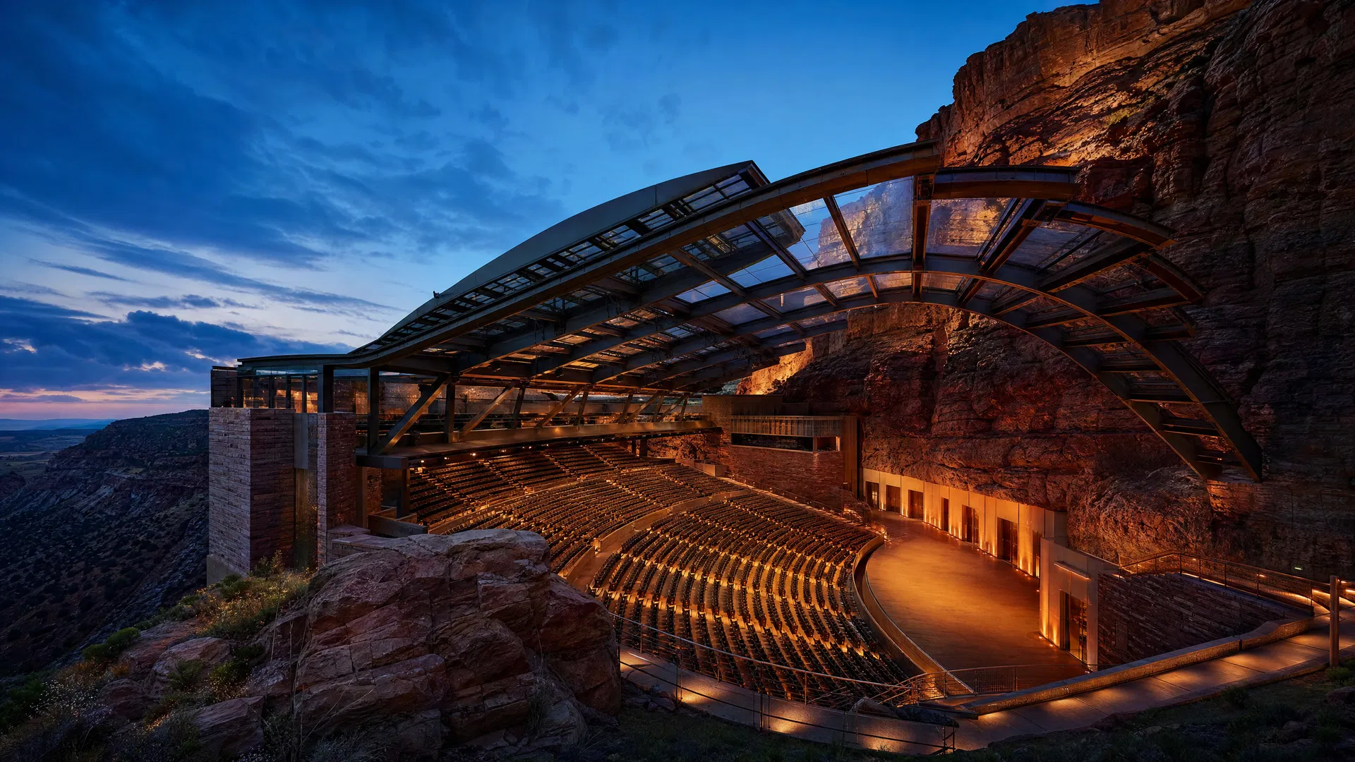 Sweeping outdoor amphitheater structure built into a canyon wall with retractable roof at twilight