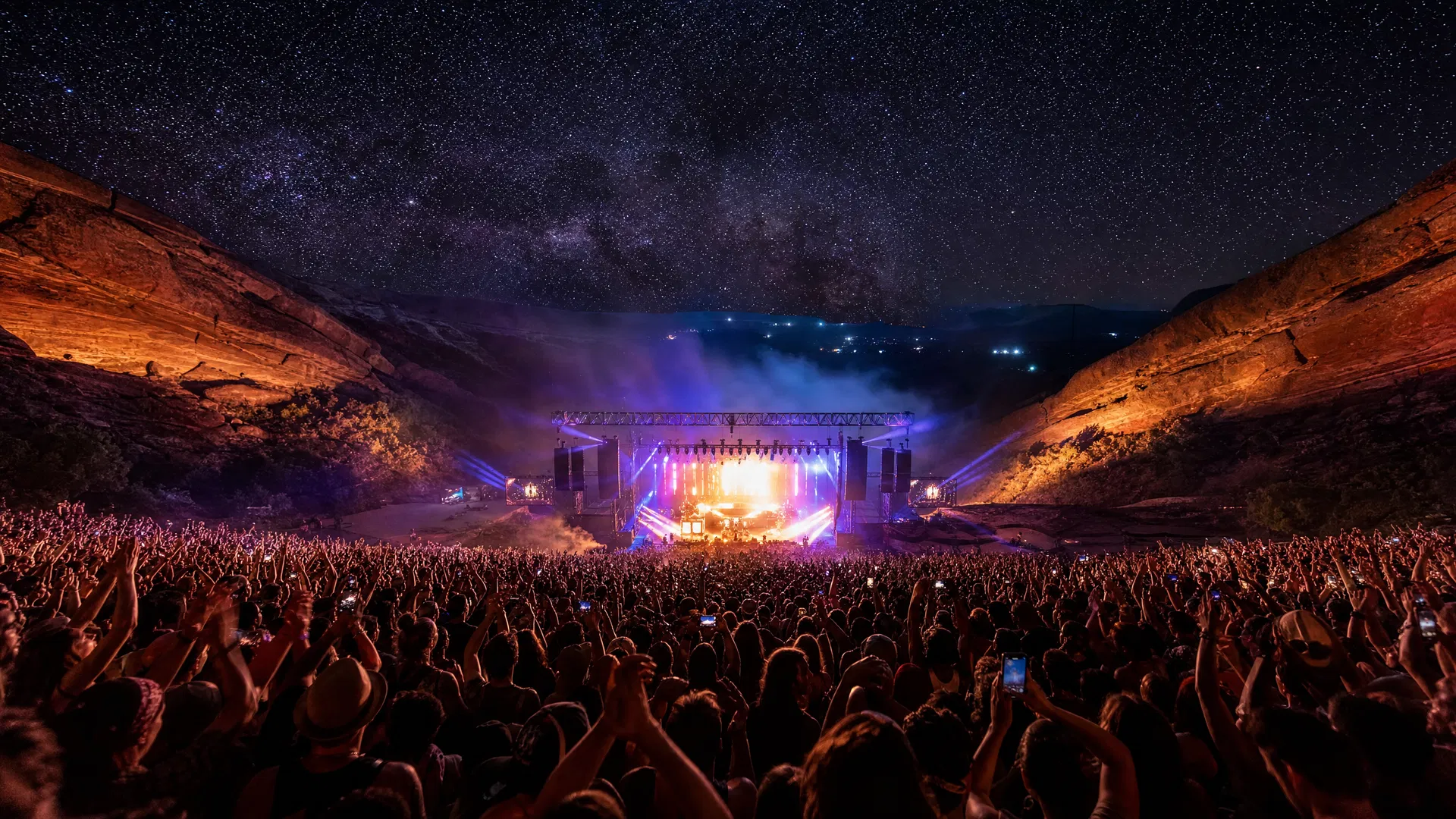 Wide-angle concert photograph from the back of an outdoor amphitheater at night with dramatic colored lights and canyon walls above