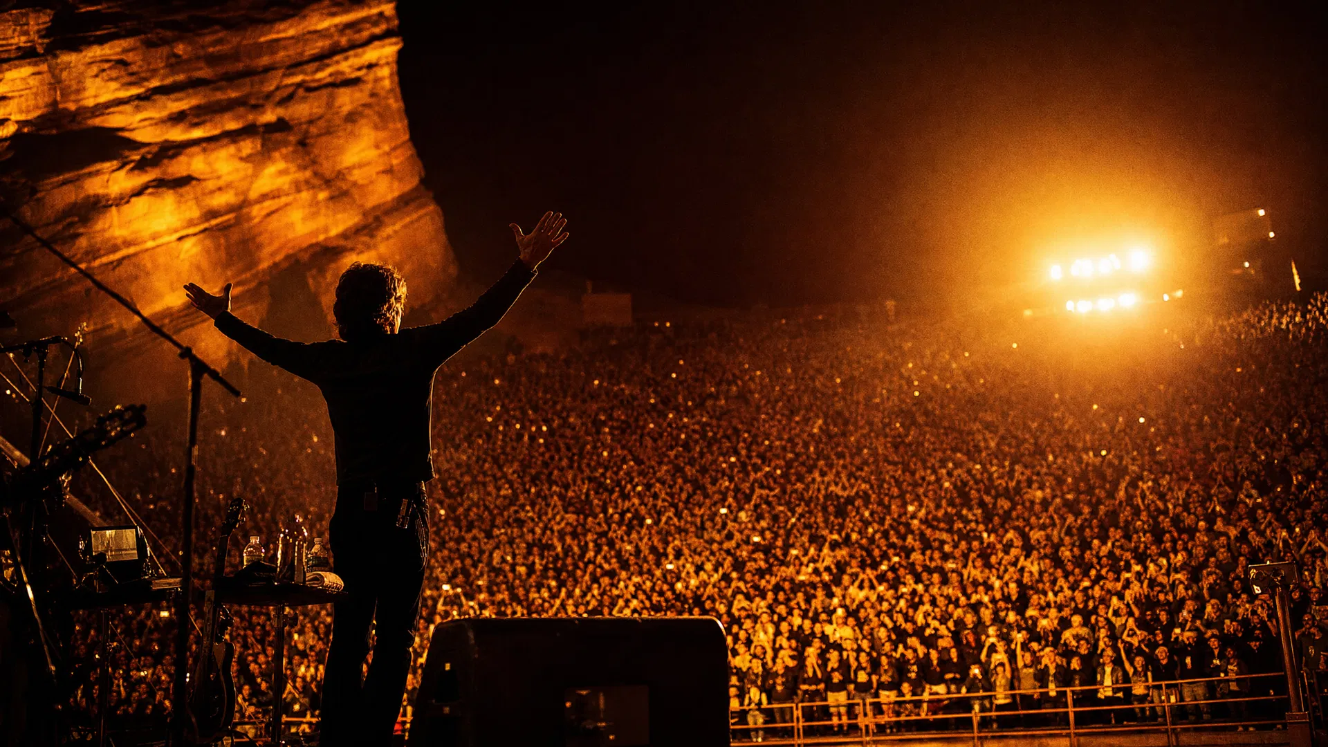 Legendary performer silhouetted against brilliant stage lights with arms raised to the crowd in a canyon amphitheater