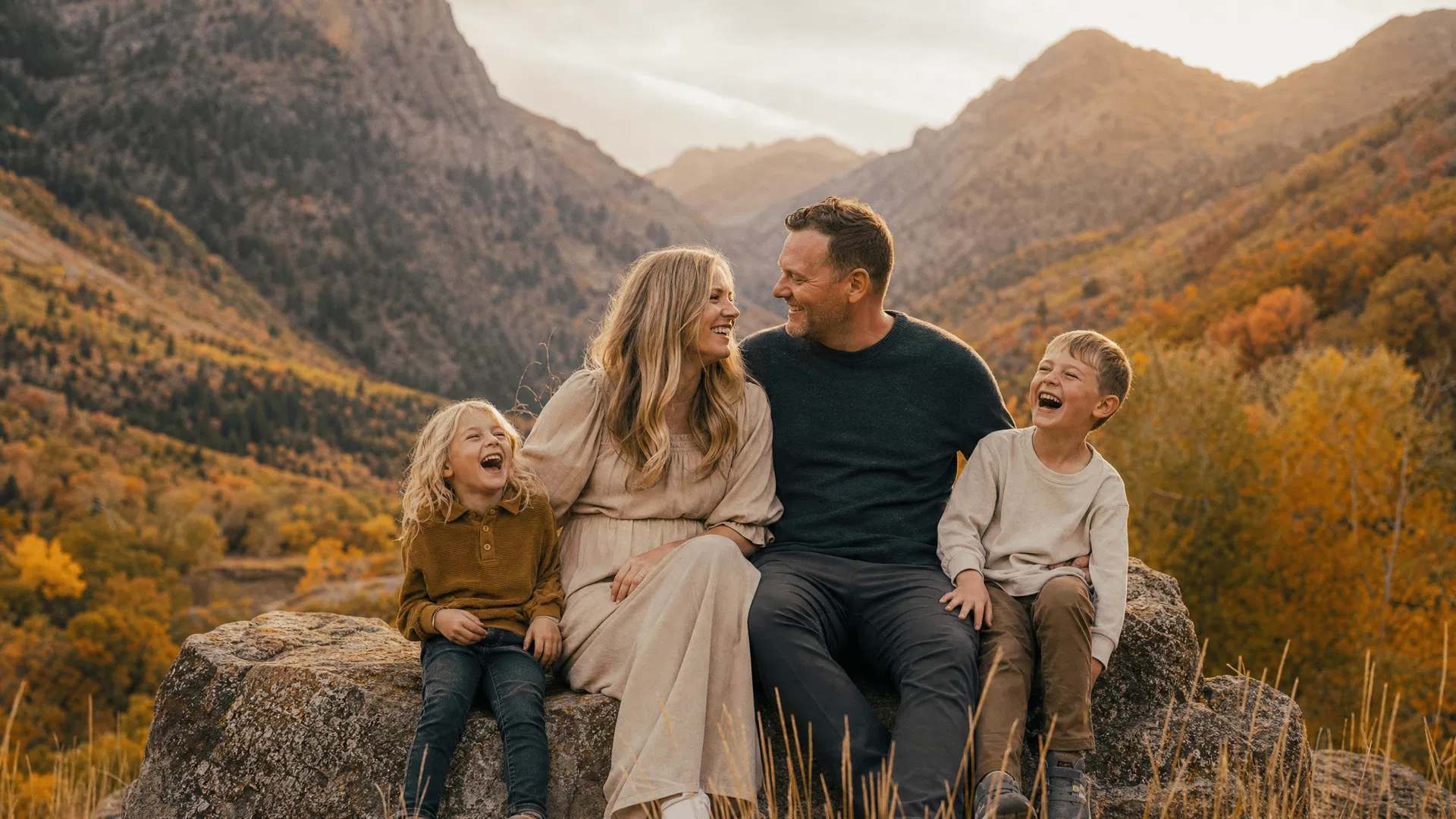 Family of four laughing together on a boulder in Provo Canyon with autumn-colored trees and canyon walls behind them