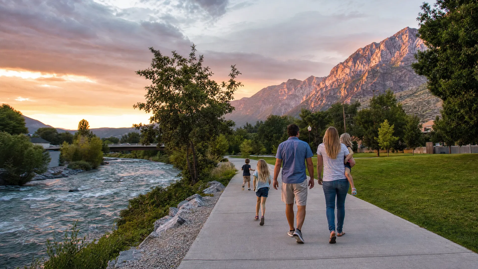 Family walking along a paved riverside trail at sunset with the Provo River and Wasatch Mountains glowing amber
