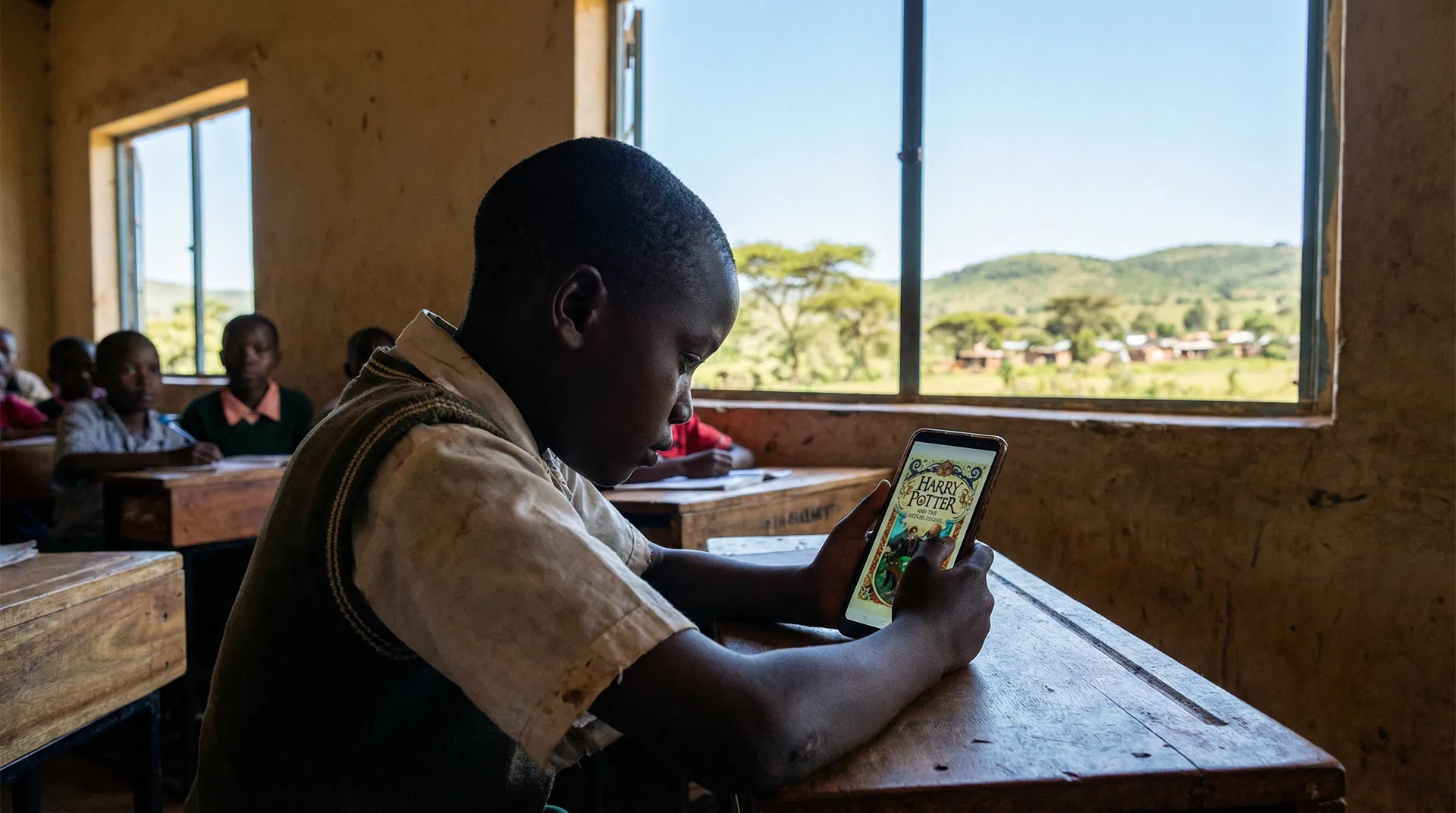 Kenyan child reading Harry Potter on smartphone in sunlit classroom with green landscape visible through window