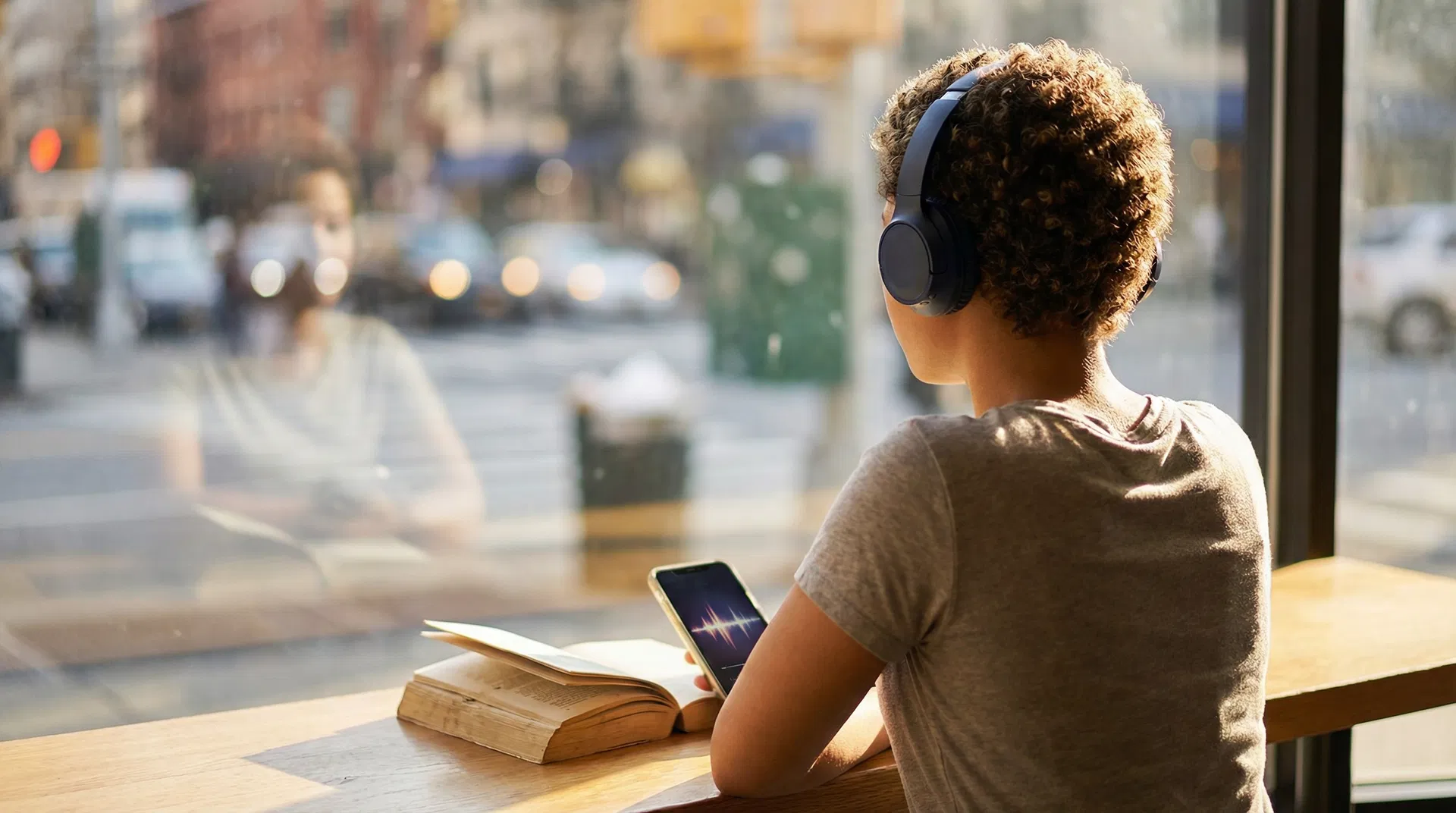 Young adult with wireless headphones in a café, open book on table beside a smartphone showing podcast waveform