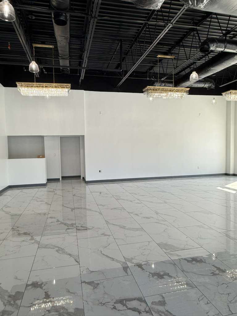 A spacious, empty event venue featuring a modern design with white walls, a black exposed ceiling, and elegant marble-patterned floor tiles. Decorative chandeliers hang from the ceiling, ready for any event setup.