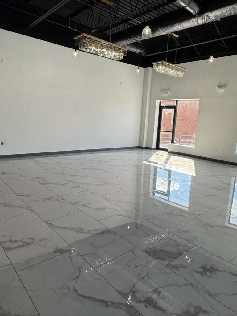An empty, modern event space featuring a glossy marble-look tile floor, white walls, exposed black ceiling, and elegant rectangular chandeliers. Natural light streams in through a large window/doorway.