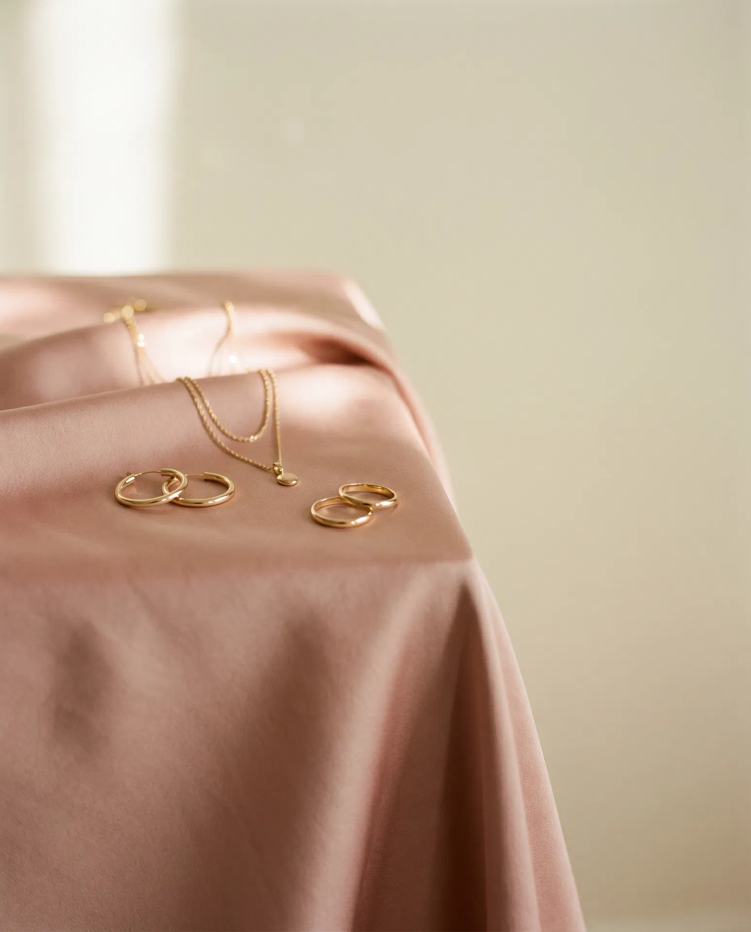 Gold­kette, Creolen und Stapelringe auf Puderrosa