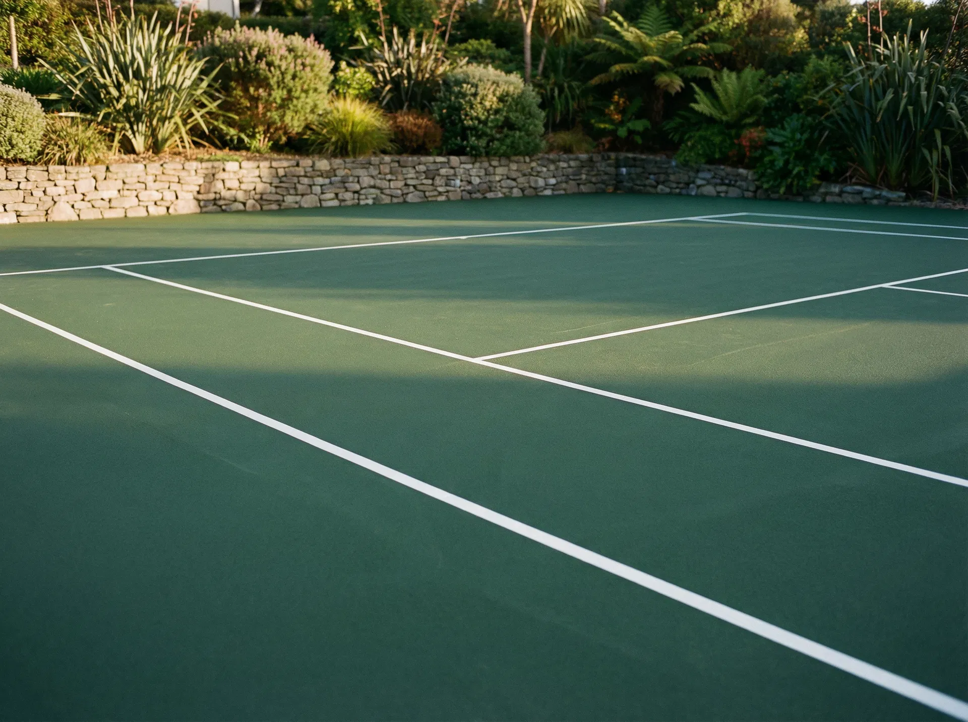 Freshly resurfaced green acrylic tennis court with crisp white lines and stone retaining wall — indicative design