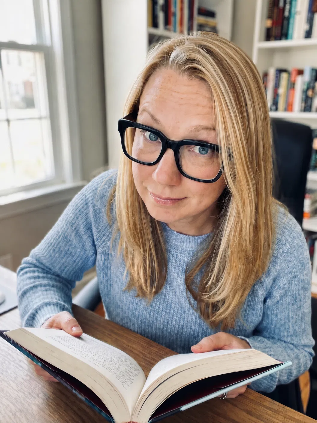 GenXFemHealth founder sitting at a desk with big black frame glasses and a light blue sweater, holding a book with a curious expression