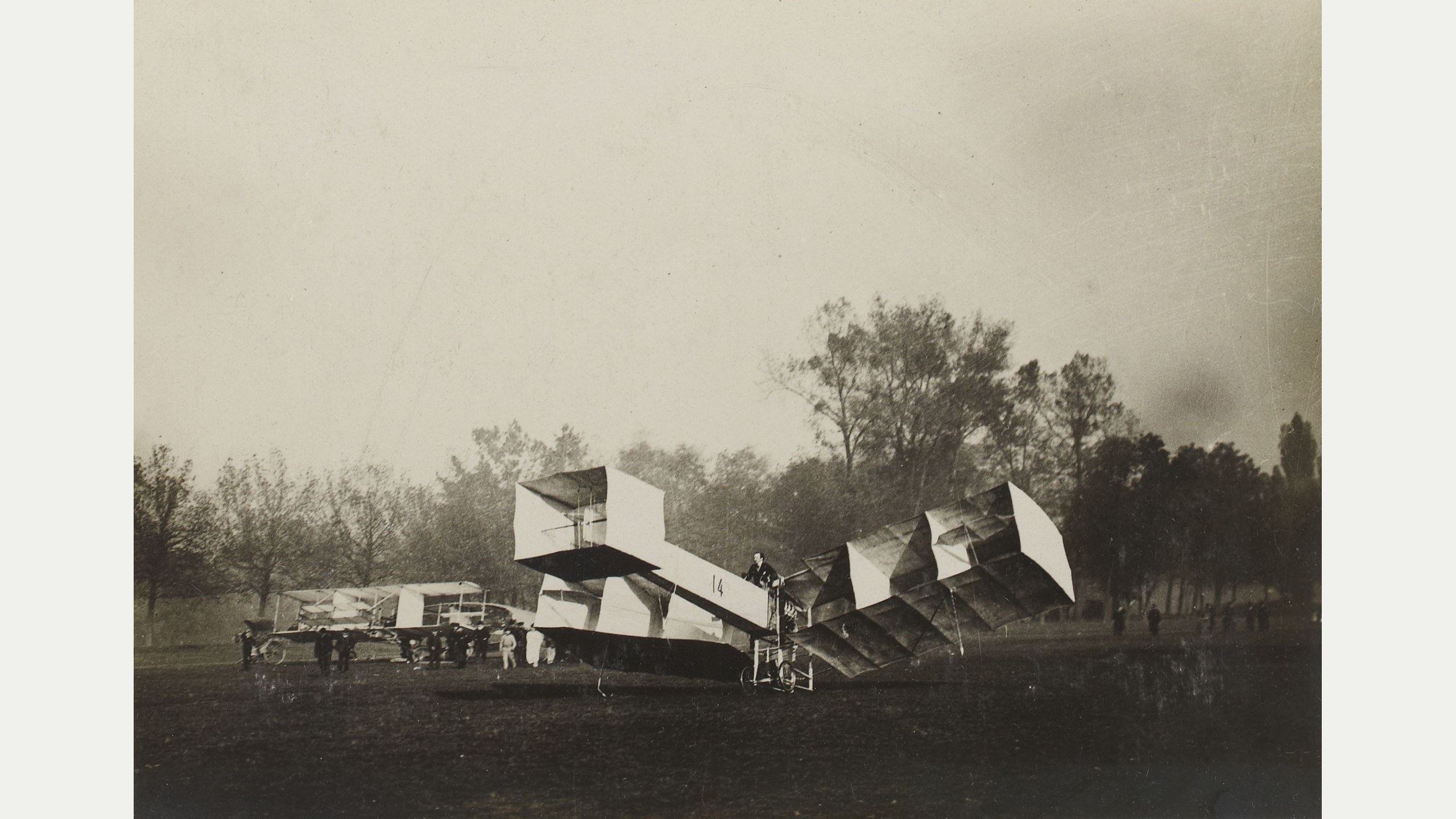 Fotografia histórica do 14-bis no solo em novembro de 1906, com Santos-Dumont na cesta, asas em forma de caixas e outro avião ao fundo.